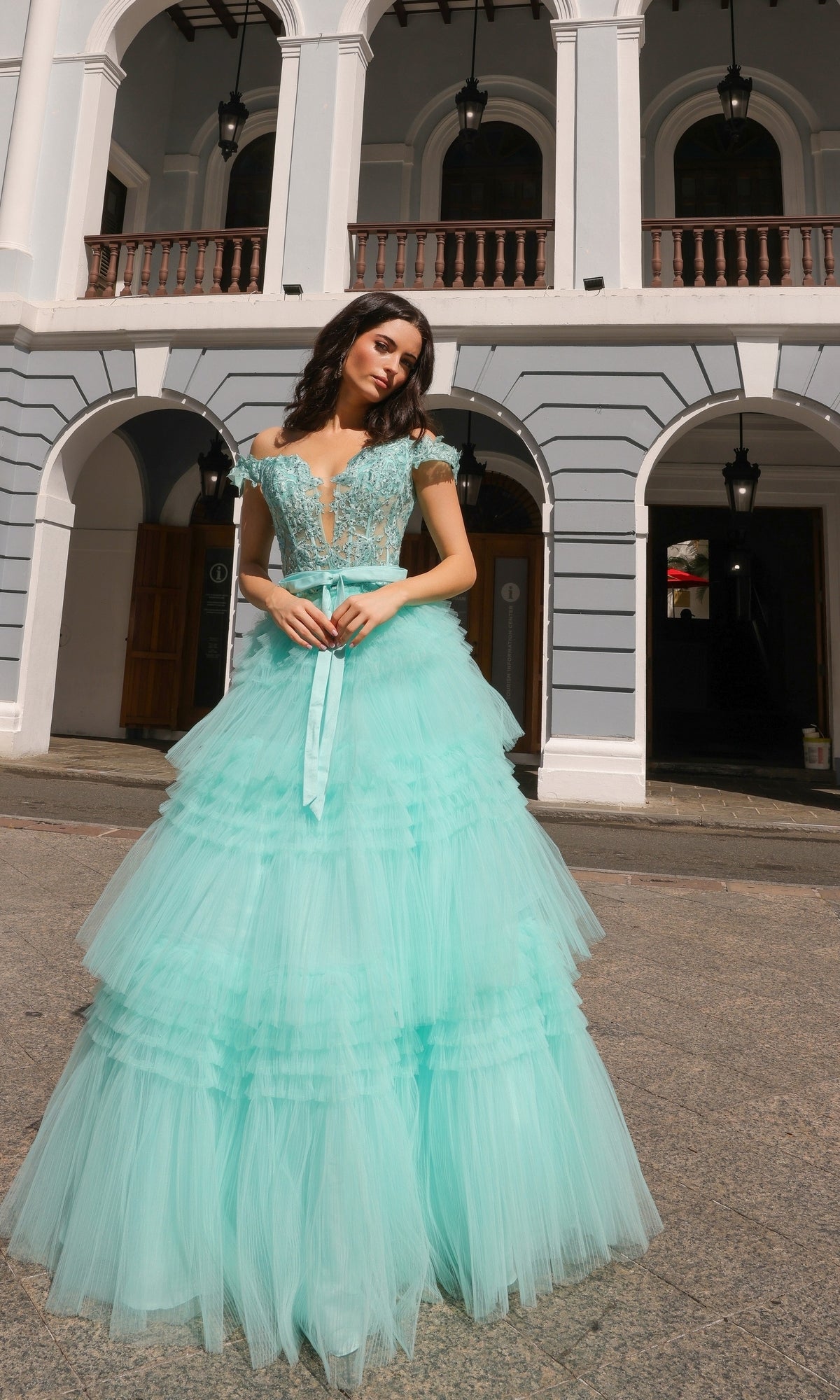 A woman stands outdoors before an arched building, wearing the Nox Anabel Designer Ruffled Prom Ball Gown E1293 featuring a layered light blue skirt and a fitted, beaded embroidered lace bodice.
